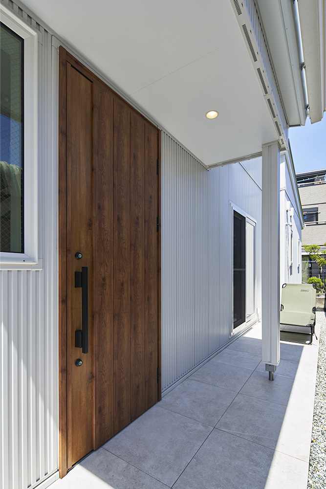 白いガルバリウムに木製ドアが映える玄関。屋根付きのアプローチで雨の日でもストレスフリー